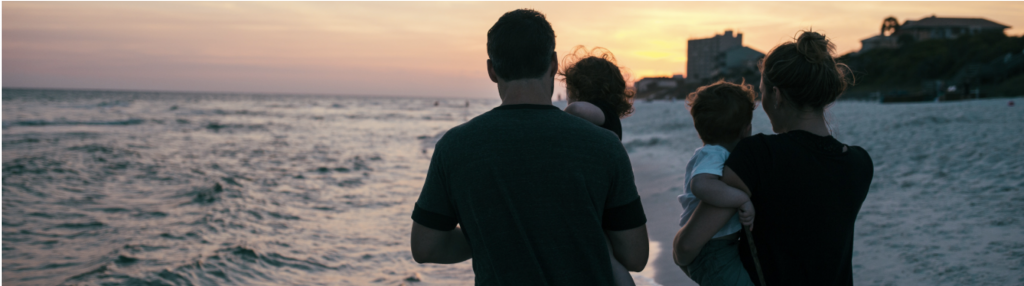 Family of four; parents carrying two young children during sunset on a beach.