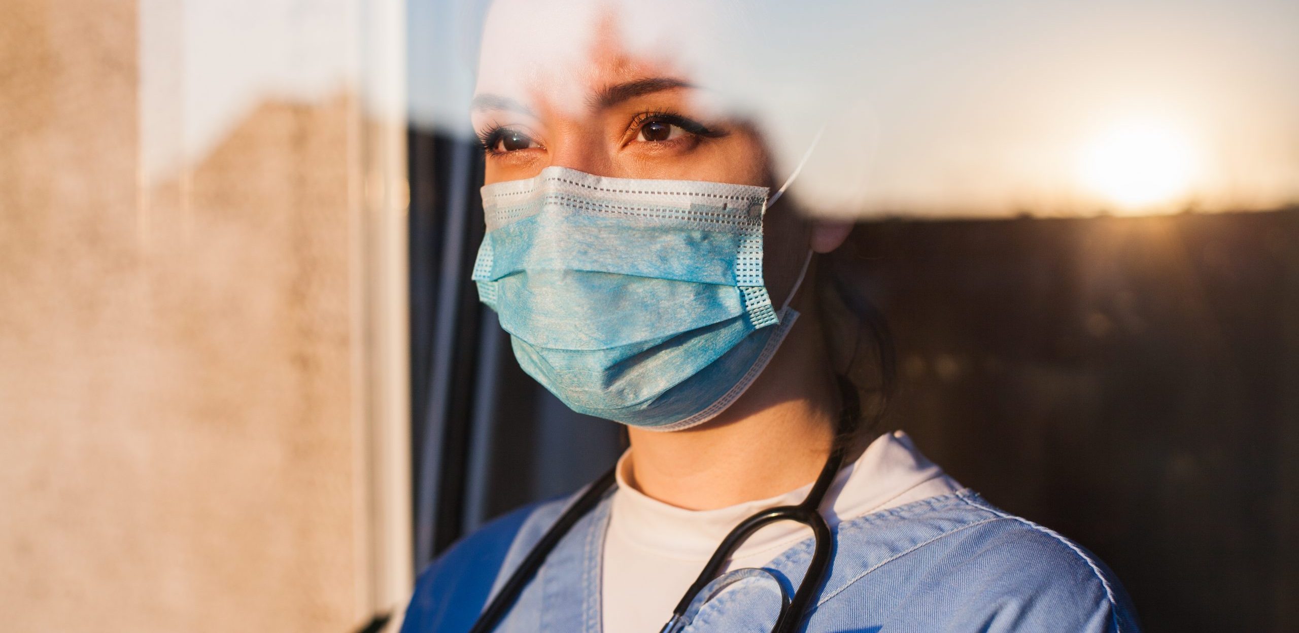 Young sad female caucasian UK US NHS EMS doctor carer looking through ICU window,fear uncertainty in eyes,wearing face mask gazing at sun,hope faith in overcoming Coronavirus COVID-19 pandemic crisis